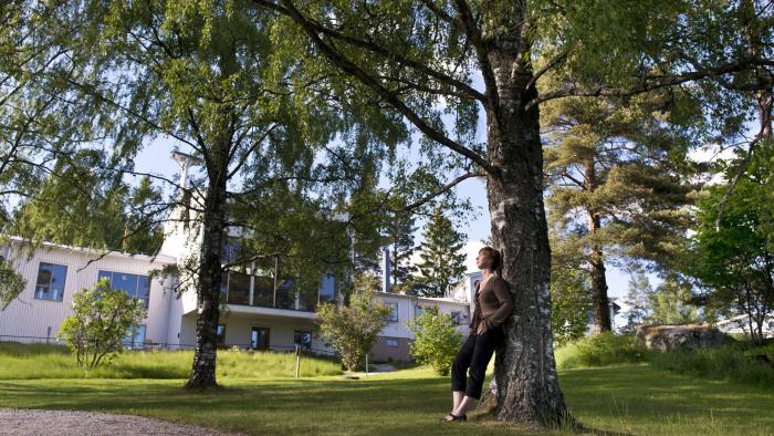 En kvinna lutar sig mot ett grönskande träd, i bakgrunden syns kursgården Solliden.