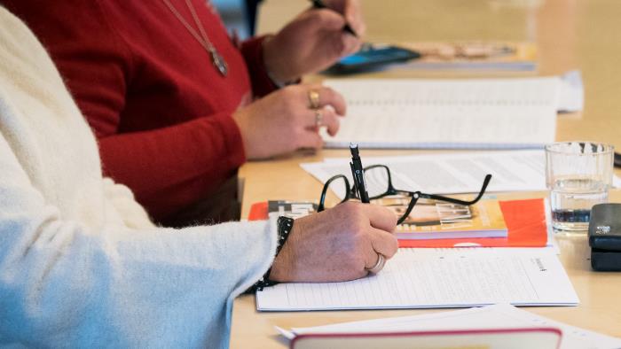 Två personer sitter på möte. På ett bord framför dem finns pennor, papper, broschyrer, glasögon, vattenglas.