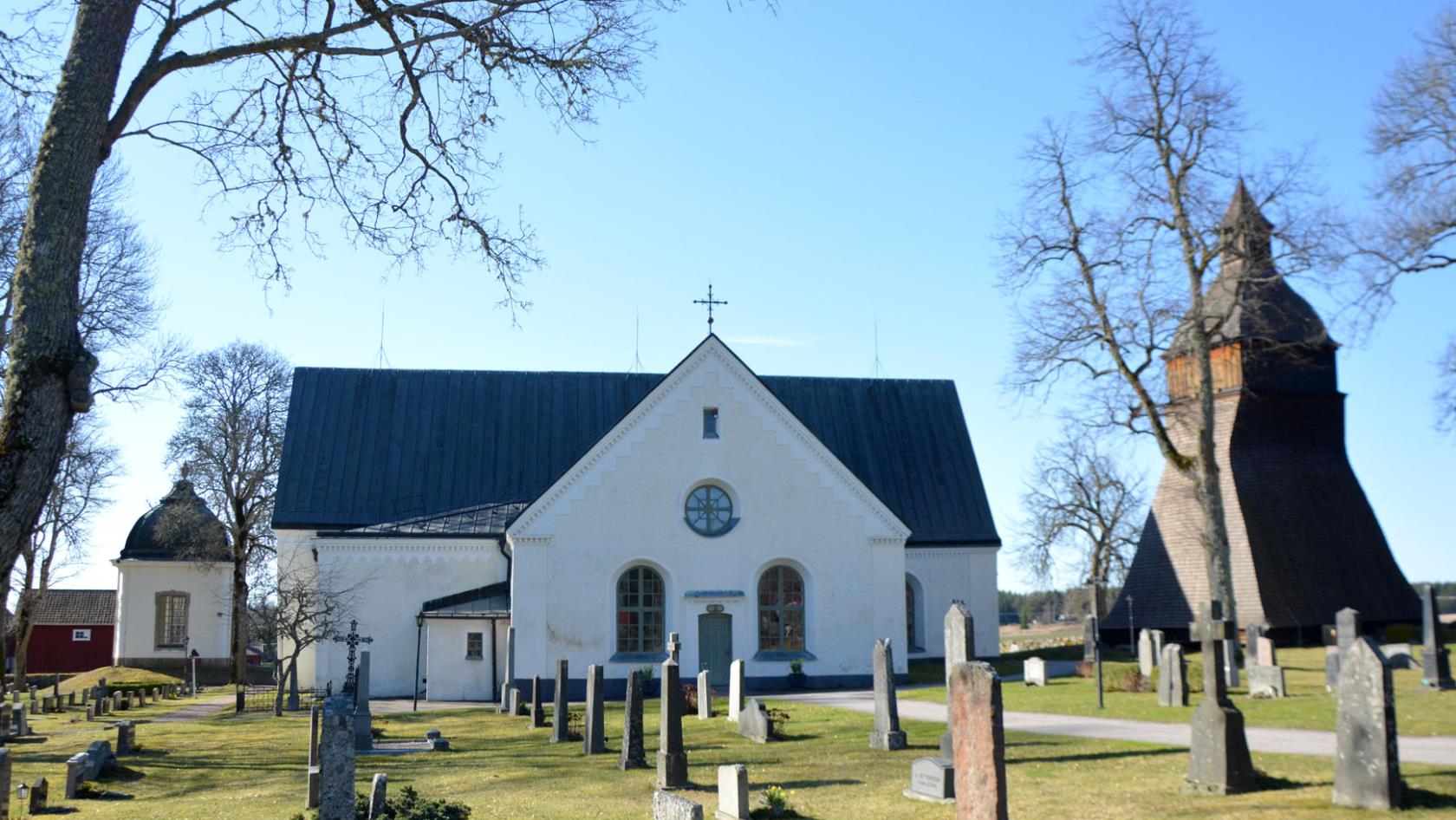 Vit stenkyrka med tjärad klockstapel till höger och litet gravkor till vänster samt gravstenar och träd i förgrunden.