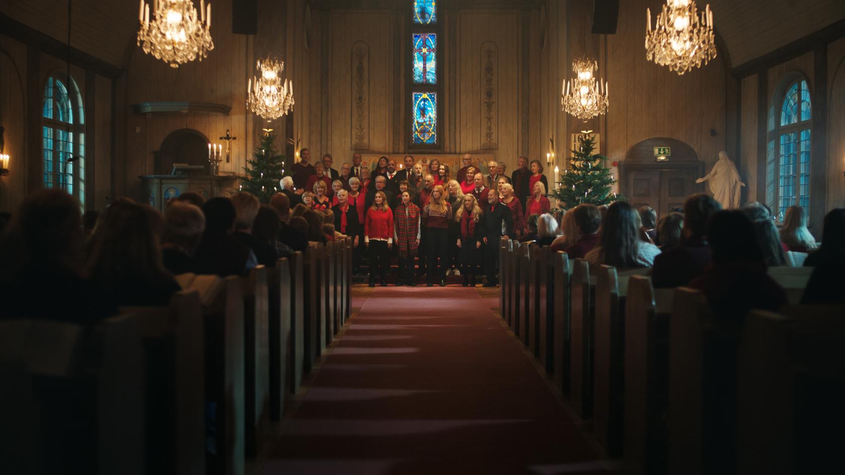 Mittgången i en fullsatt kyrka. Längst fram står en kyrkokör i julkläder mellan två julgranar och sjunger.